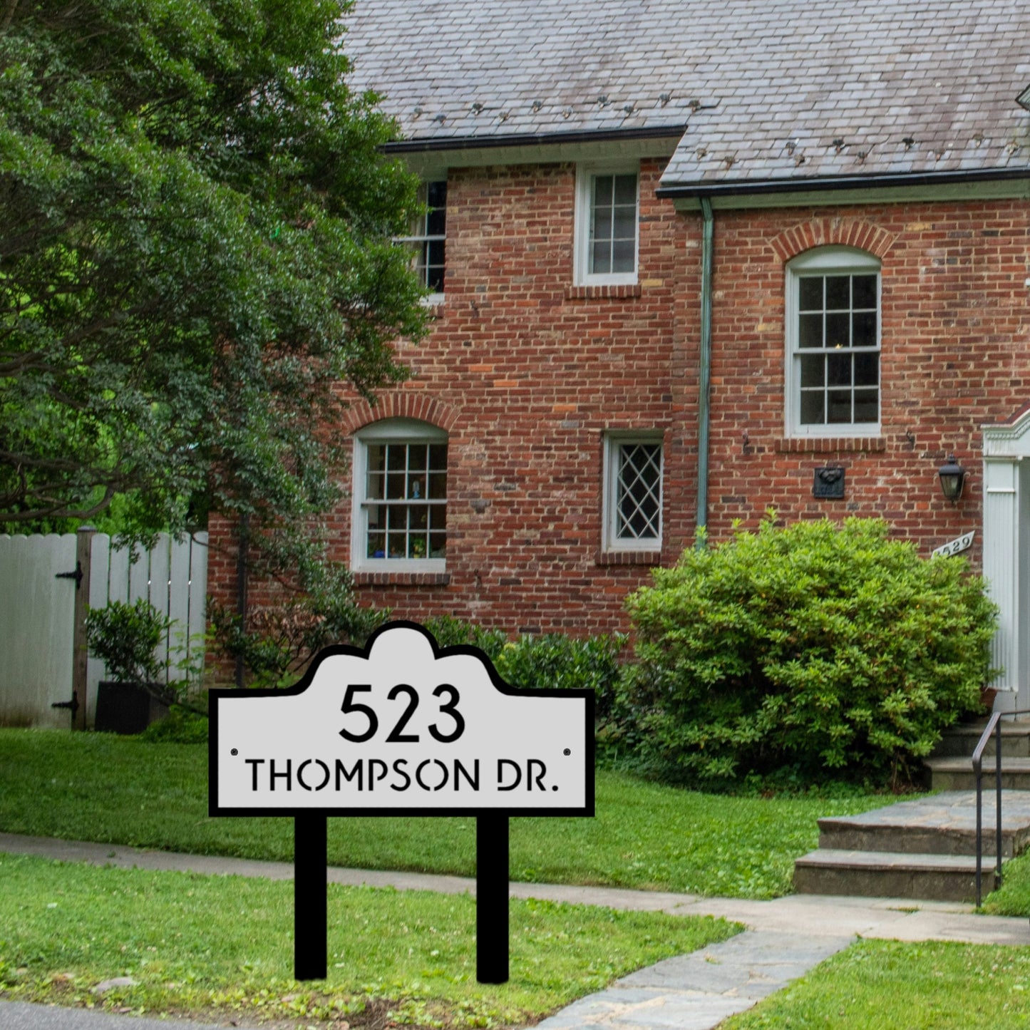 The Double Layer Luxury Address Yard Sign is elegantly positioned in the front yard of a charming classic red-brick home, exuding historic appeal. The sign showcases a sharp and tidy design, featuring a white front panel bordered in bold black. This combination creates a striking and sophisticated contrast that guarantees excellent visibility.