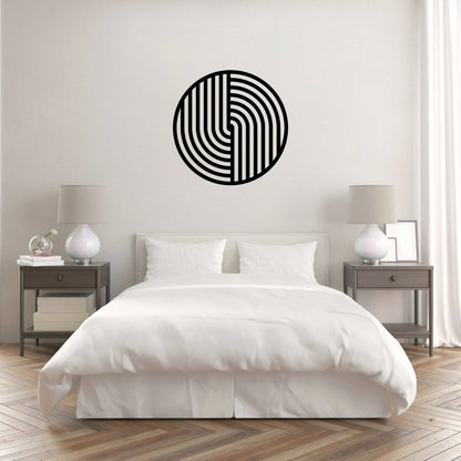 The image shows a modern bedroom with a minimalist design. A round metal wall art piece with a geometric black pattern hangs above the bed, creating a striking contrast against the light-colored wall. The decor gives a clean, contemporary, and sophisticated feel.