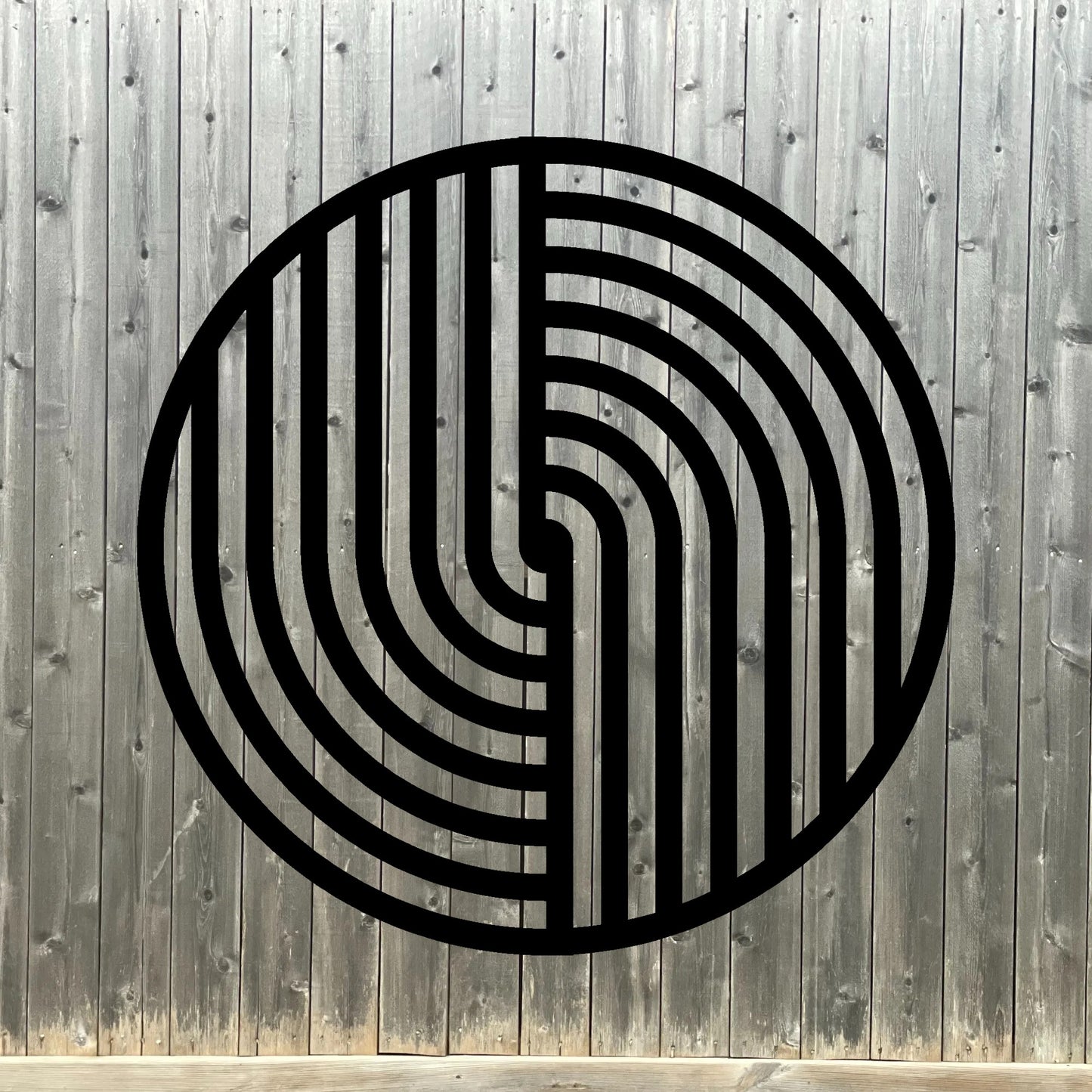 The image features a round metal wall art piece with a bold black geometric pattern, mounted on a dark wooden fence background. The contrast between the deep wood tones and the crisp black lines of the artwork creates a striking, modern aesthetic with a touch of rustic charm.