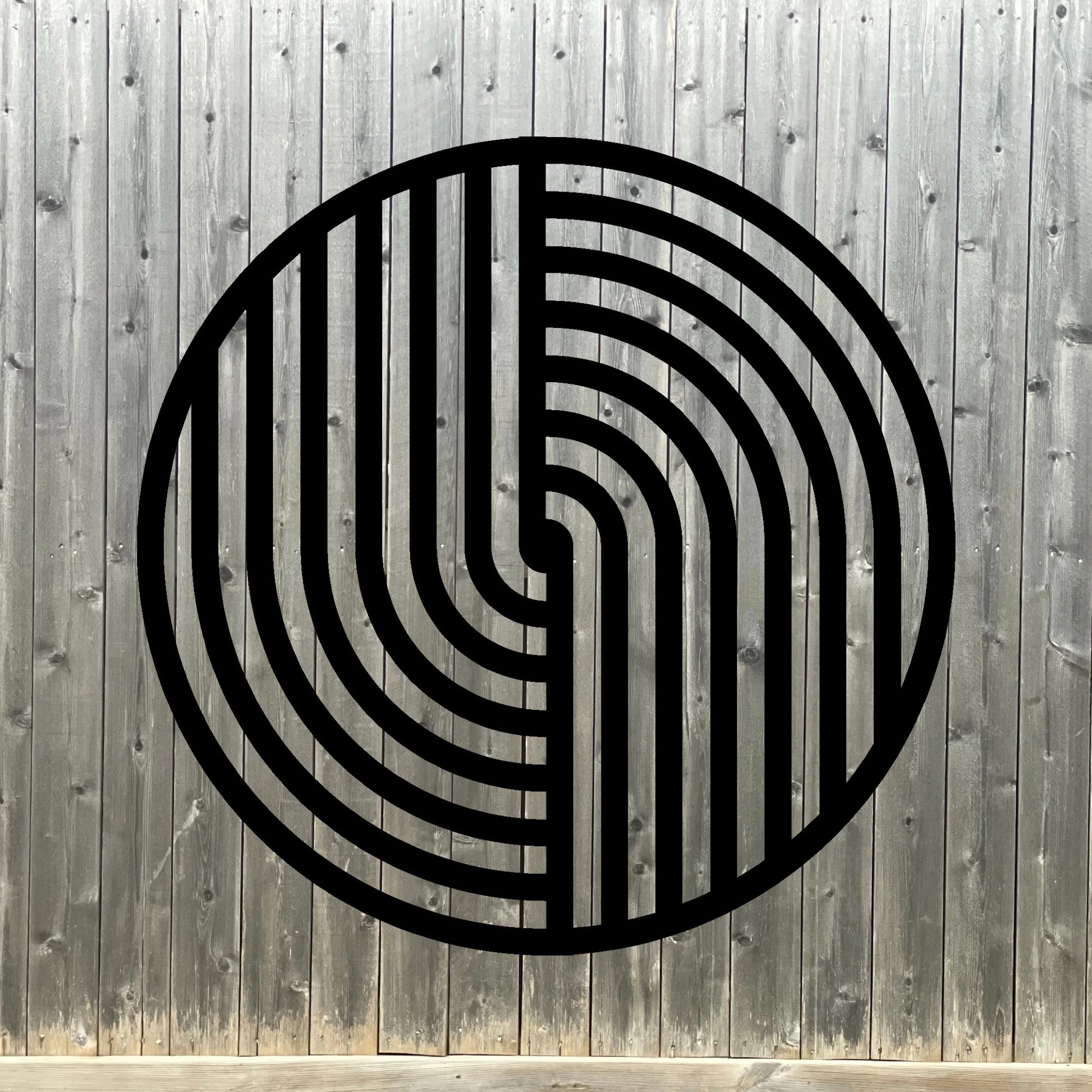 The image features a round metal wall art piece with a bold black geometric pattern, mounted on a dark wooden fence background. The contrast between the deep wood tones and the crisp black lines of the artwork creates a striking, modern aesthetic with a touch of rustic charm.