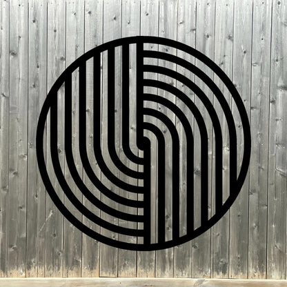 The image features a round metal wall art piece with a bold black geometric pattern, mounted on a dark wooden fence background. The contrast between the deep wood tones and the crisp black lines of the artwork creates a striking, modern aesthetic with a touch of rustic charm.