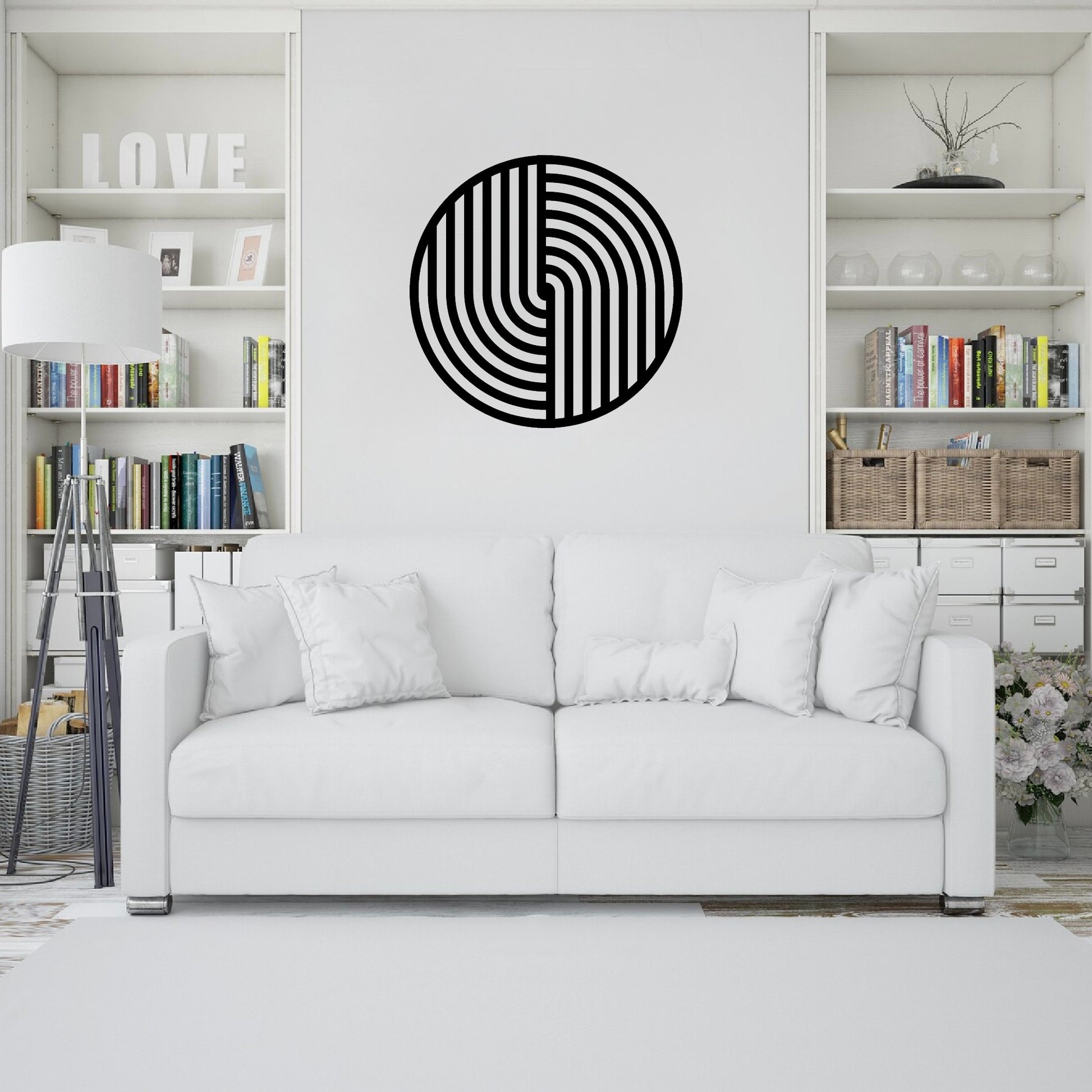 The image showcases a modern living room with a minimalist design. A round metal wall art piece with a bold geometric black pattern hangs above a white sofa, adding a striking contrast to the neutral decor.