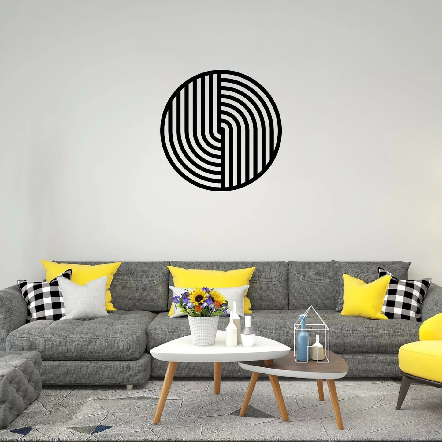 The image depicts a stylish and contemporary living room featuring a round metal wall art piece with a bold black geometric design hanging above a gray sectional sofa. The room has a modern color scheme with pops of yellow and black-and-white checkered pillows.