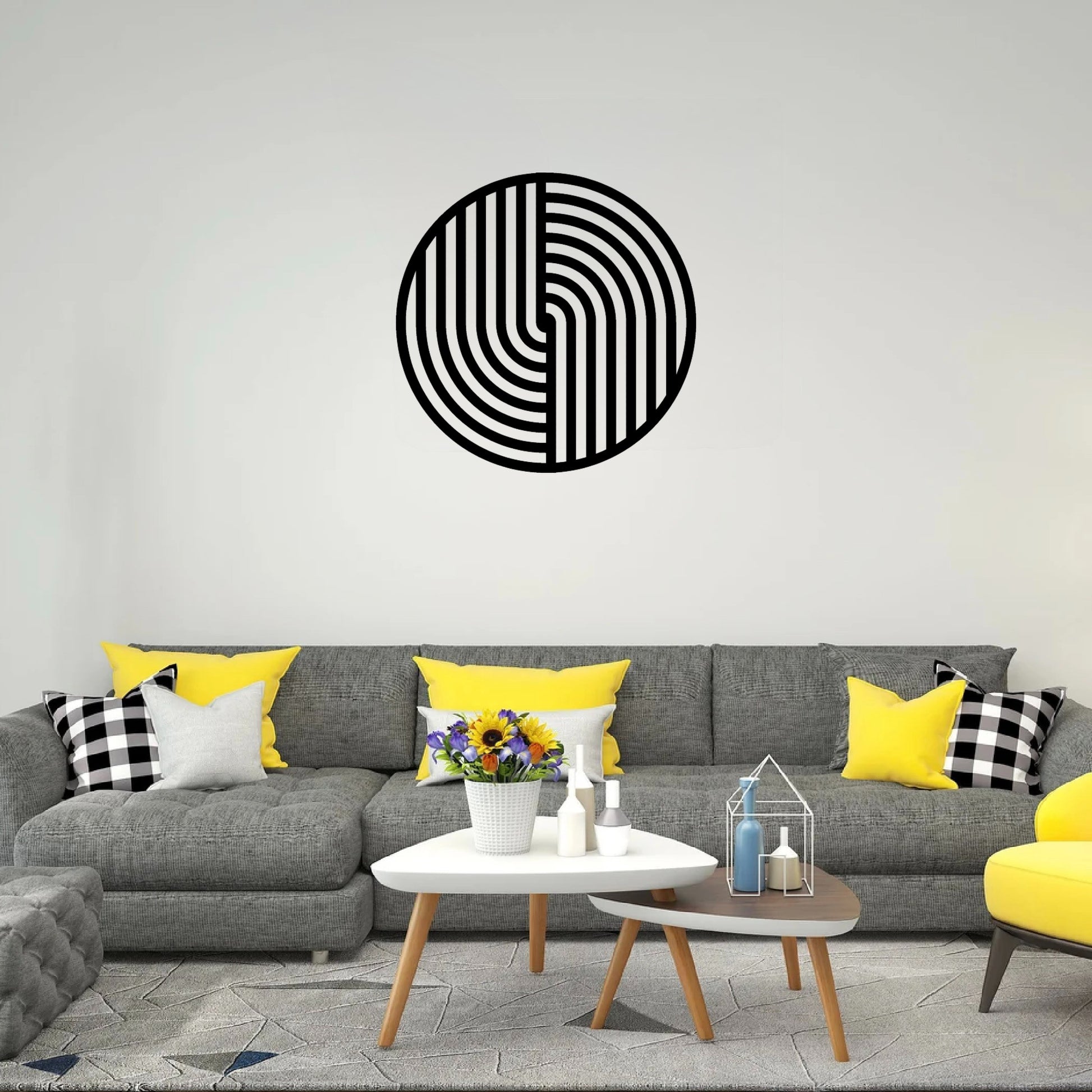 The image depicts a stylish and contemporary living room featuring a round metal wall art piece with a bold black geometric design hanging above a gray sectional sofa. The room has a modern color scheme with pops of yellow and black-and-white checkered pillows.
