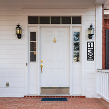 Load image into Gallery viewer, Front door of a white house with a white door and a black vertical home numbers sign hanging to the right of the door